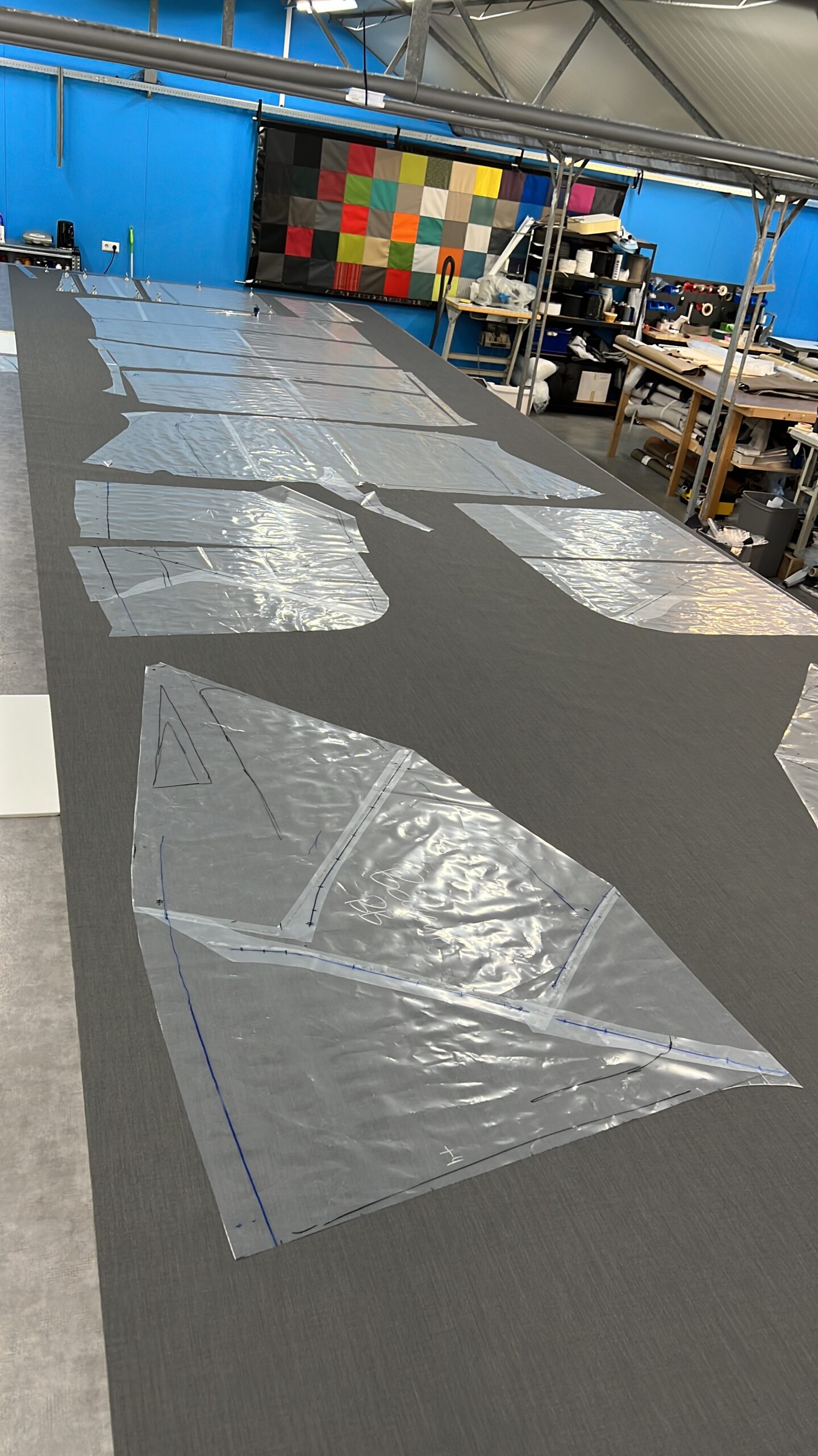 Long grey worktable covered with large sheets of silver material taped in place, in a bright workshop with blue walls and storage shelves in the background.