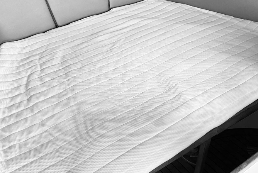 Quilted white mattress protector on a bed with a cushioned headboard visible at the top left, simple, clean bedding look.