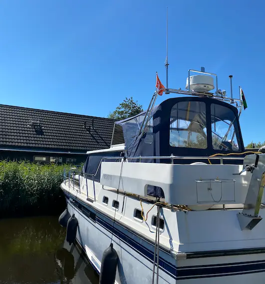 White motor yacht moored along a canal, with a blue canvas cabin, fenders along the hull, flags at the stern, and a house with a tiled roof in the background under a blue sky.