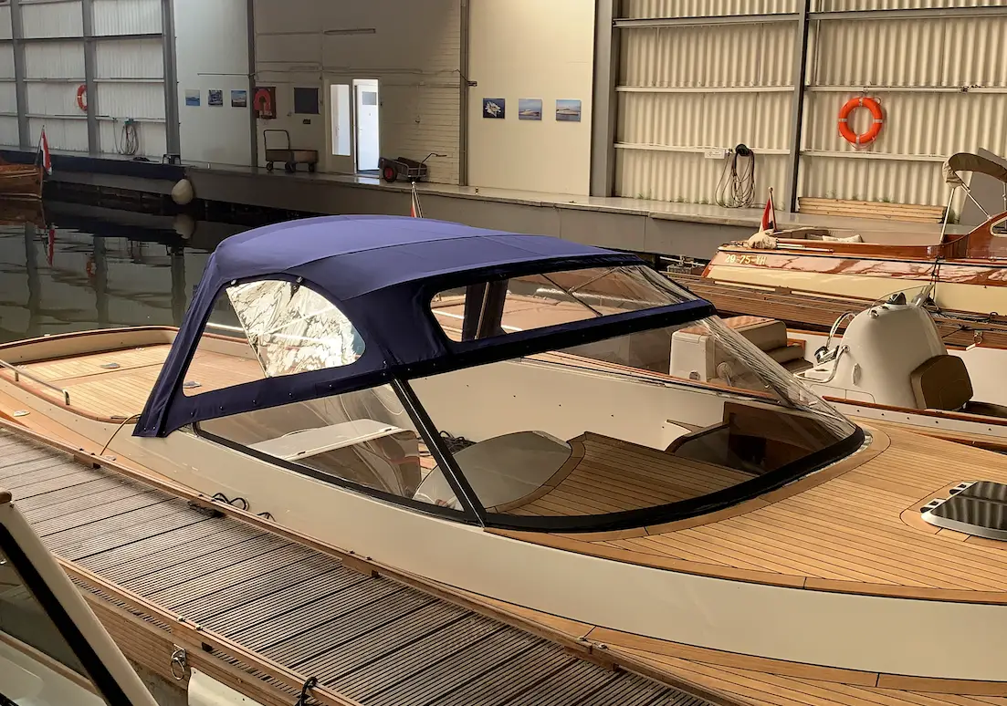 A white luxury powerboat with a blue canvas cabin cover docked indoors, wooden deck and other boats nearby in a marina shed.