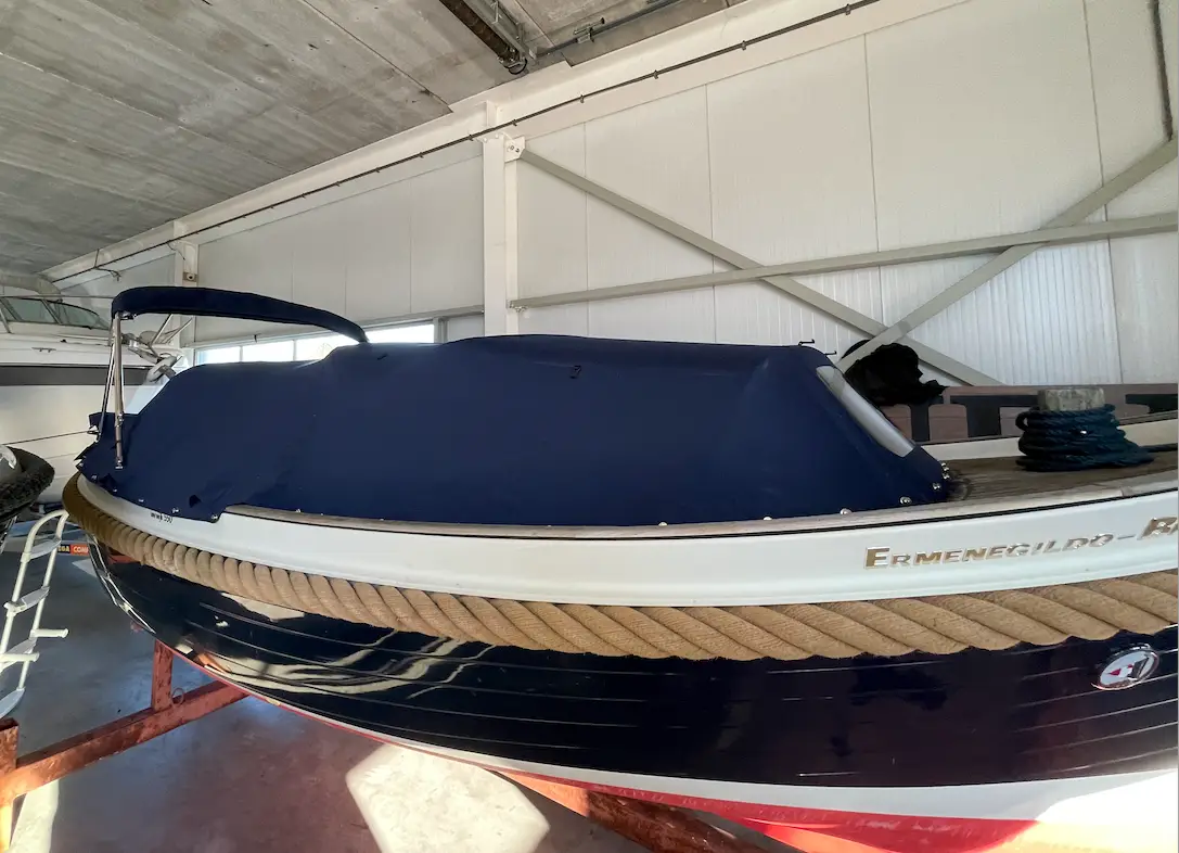 Blue-covered speedboat sits on a trailer inside a metal-boathouse, rope fenders wrapped around the white and dark hull edge.