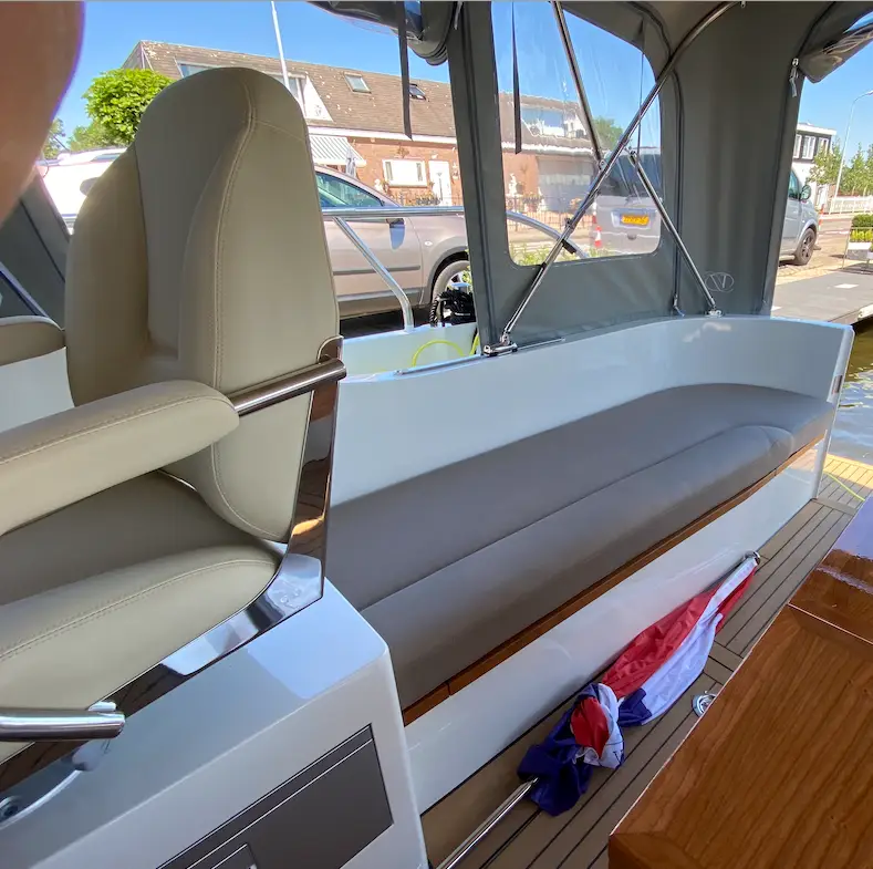 Inside a small motorboat: long beige cushioned bench, cream captain seat, wood trim, and gray canopy with clear side windows.
