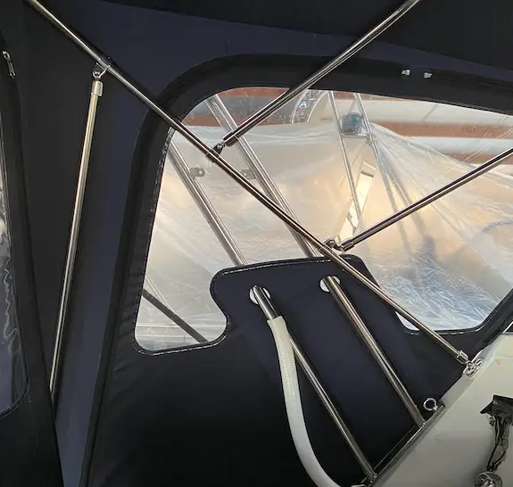 Inside a sailboat cockpit showing metal support poles, navy canvas, and a white hose attached to a frame with a plastic window in the background