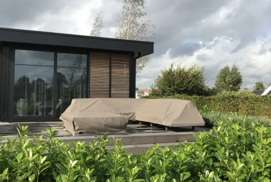 Modern house with glass doors; beige-covered outdoor furniture on a wooden deck amid hedges and green plants.