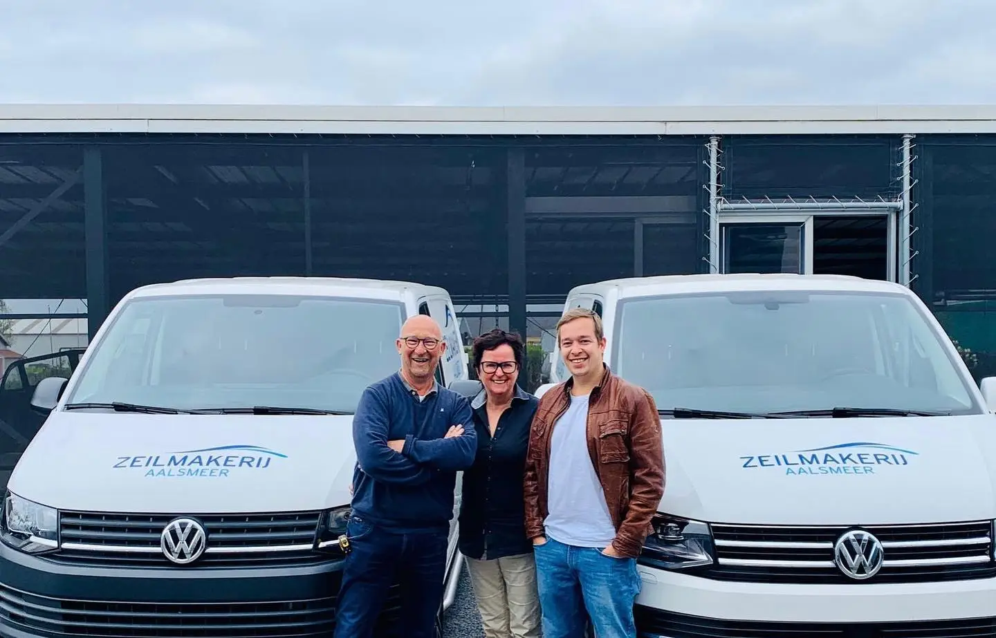 Three adults pose smiling in front of two white Volkswagen vans labeled Zeilmakerij Aalsmeer outside a modern building.