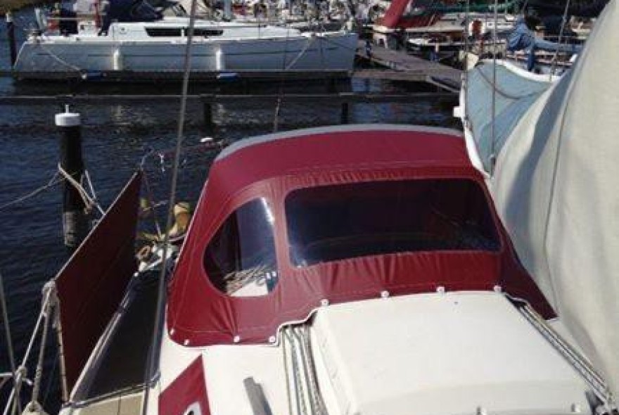 Red-canvas-covered cockpit of a small sailboat docked at a marina, with other boats and calm water in the background.