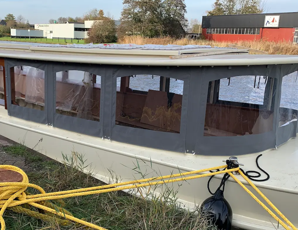 Boat with a gray canvas canopy tied to a docked mooring, by calm water and grassy riverbank.