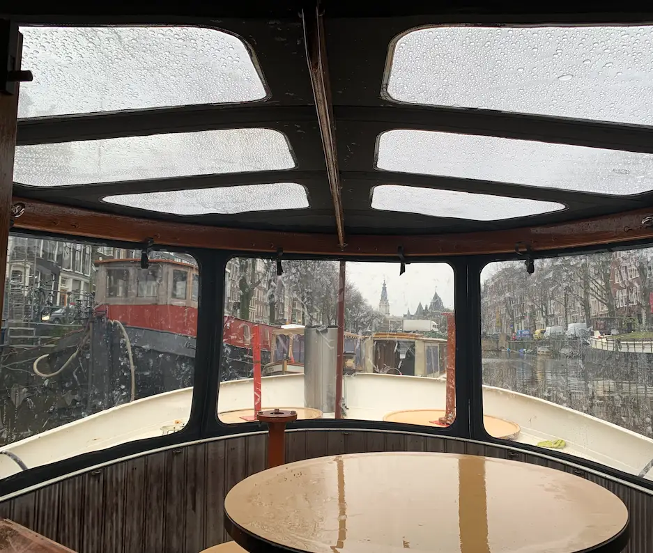 Interior of a small boat cabin with a glossy circular wooden table, rain-streaked windows, and a canal street scene visible ahead.