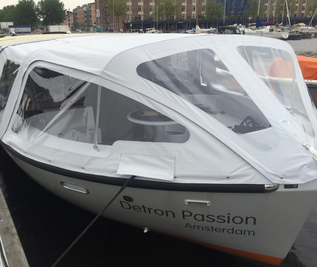 White motorboat with a soft white canopy docked at a marina; hull shows the name “Detron Passion” and port of Amsterdam.