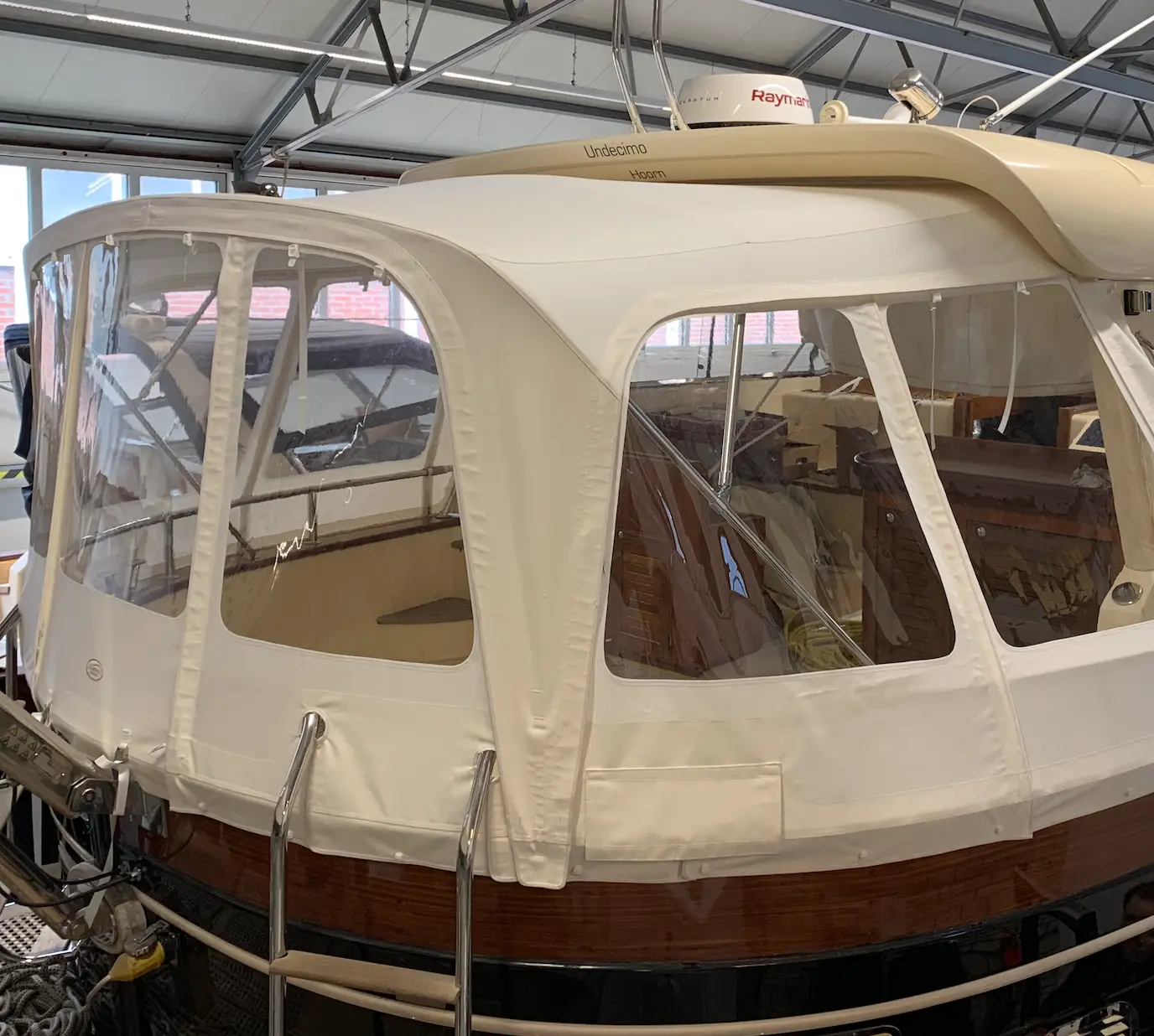 White canvas boat enclosure with clear plastic windows on a wooden-hulled small yacht inside a workshop.