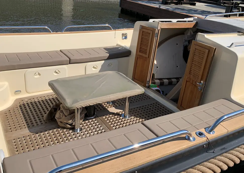 Open yacht cockpit with a small rectangular table on metal legs, cushioned bench seating, and wooden cabin doors ajar exposing the interior.