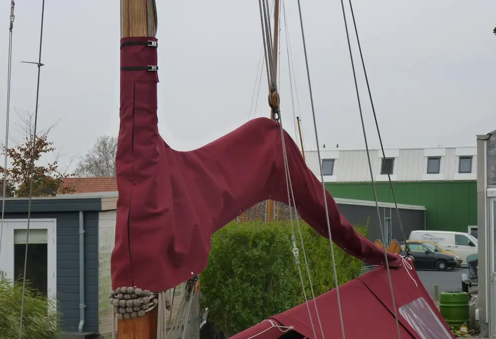 Red sail cover wrapped around a sailboat's mast in a boatyard, with rigging and a green building in the background.