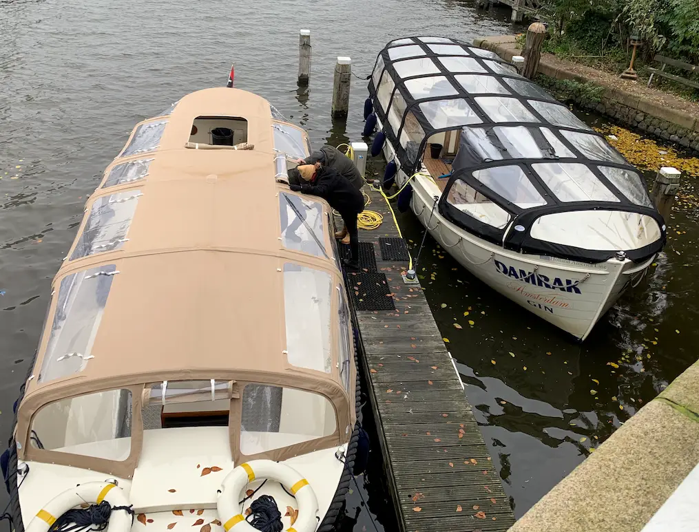 Two covered passenger boats are docked side by side at a wooden pier on the water; a person leans over the beige boat inspecting or working on its deck while a yellow hose sits nearby.