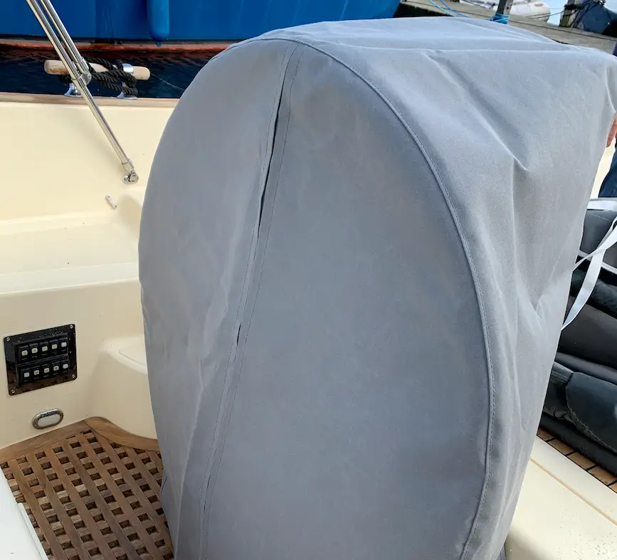 Gray protective cover over a boat's outboard motor on a wooden deck, with blue hull and dock gear in the background as seen from the stern.