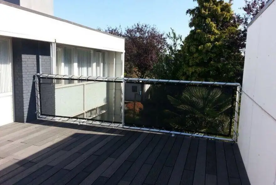Outdoor wooden deck with a metal-framed railing featuring glass panels and partial shade from the building wall beside it