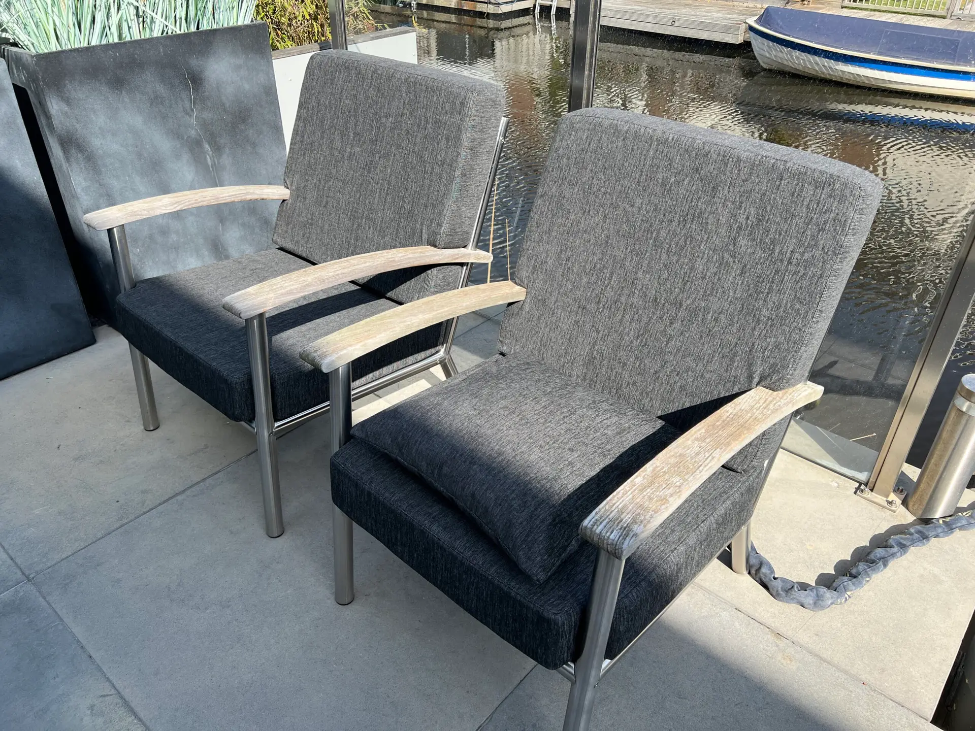 Outdoor seating: three gray upholstered chairs with light wood armrests on a concrete deck beside a canal, with a blue-and-white boat tied up in the water behind them.