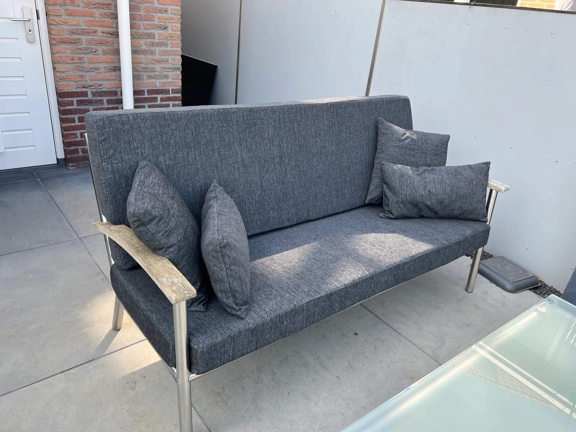 Outdoor grey fabric sofa with cushions on a patio against a brick wall and grey panels behind it.