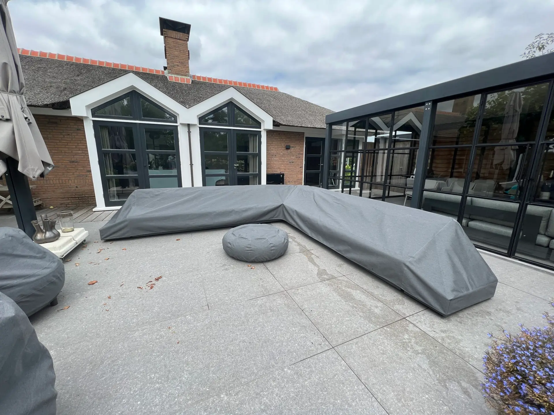 Covered outdoor furniture on a modern patio; brick house with glass-walled extension in the background.