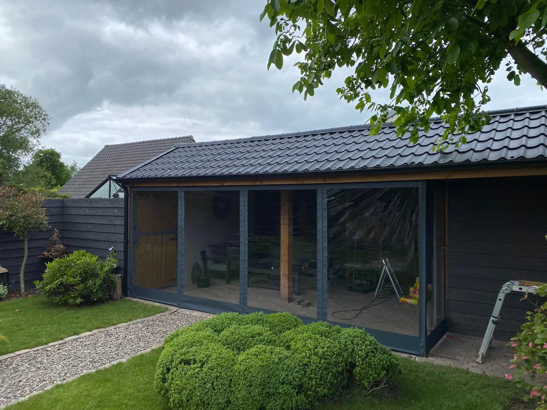 Backyard with a glass conservatory, dark-framed doors, and a tiled roof, surrounded by green shrubs and a gravel path. (Informative)