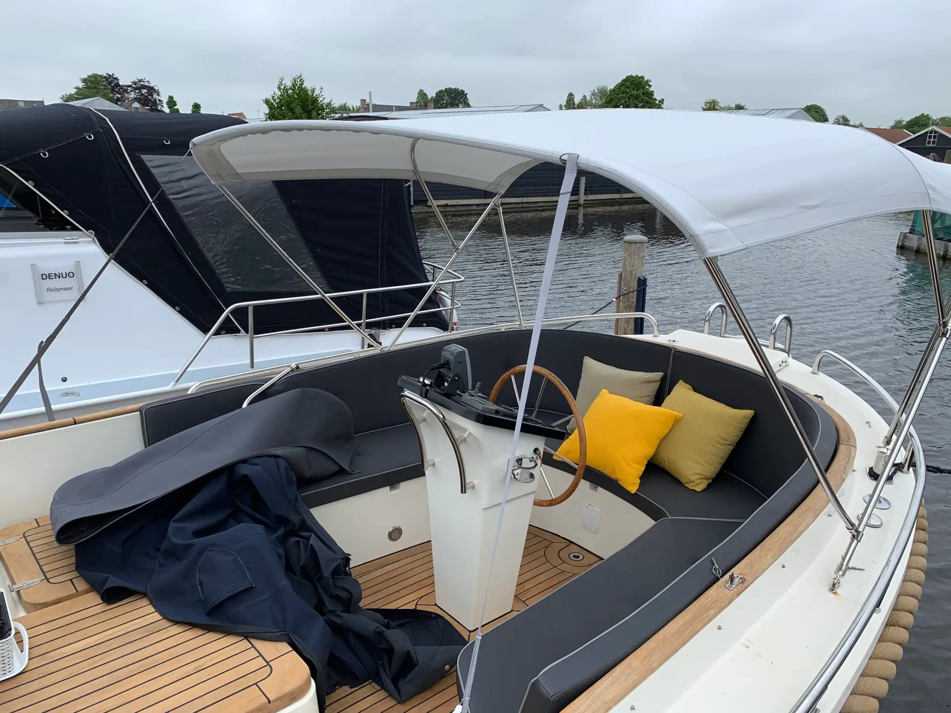 Cockpit of a white motor yacht with navy cushions and yellow accent pillows on a teak deck, moored by calm water.