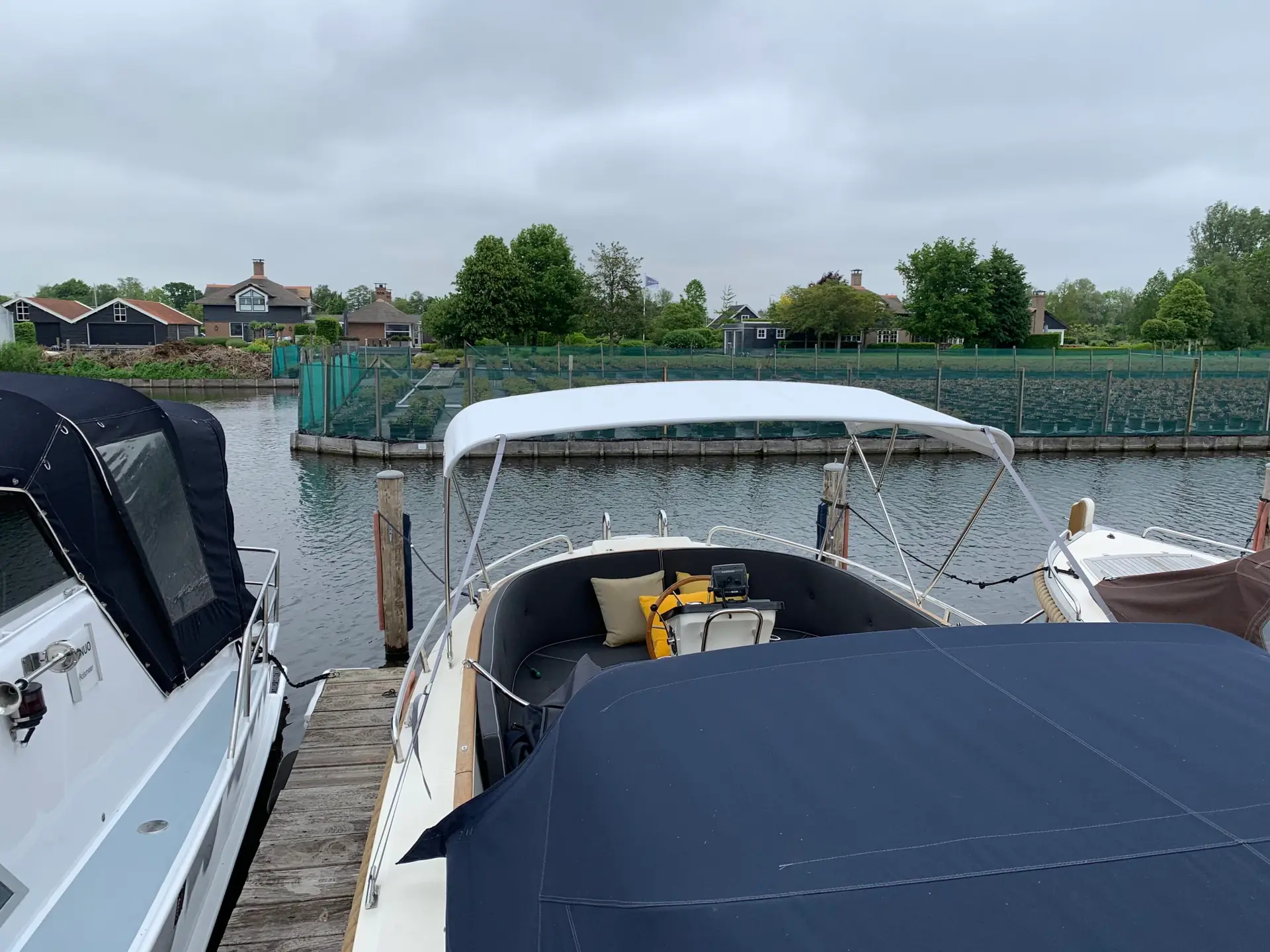 Boats docked along a wooden pier at a calm marina with houses and trees in the background under a cloudy sky.