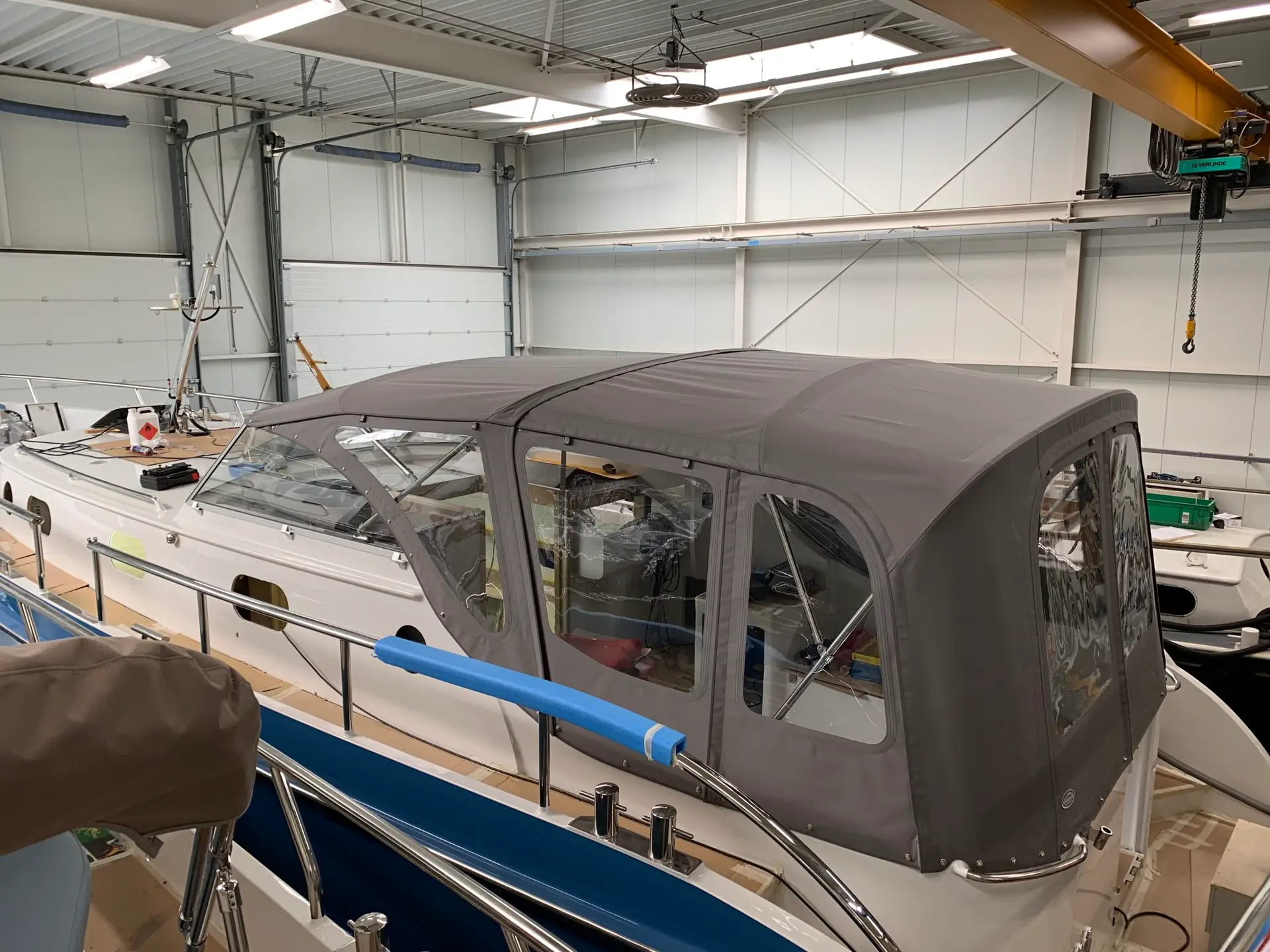 Indoor boat workshop: a gray-canvas covered boat inside a large warehouse with a yellow overhead crane in the background.